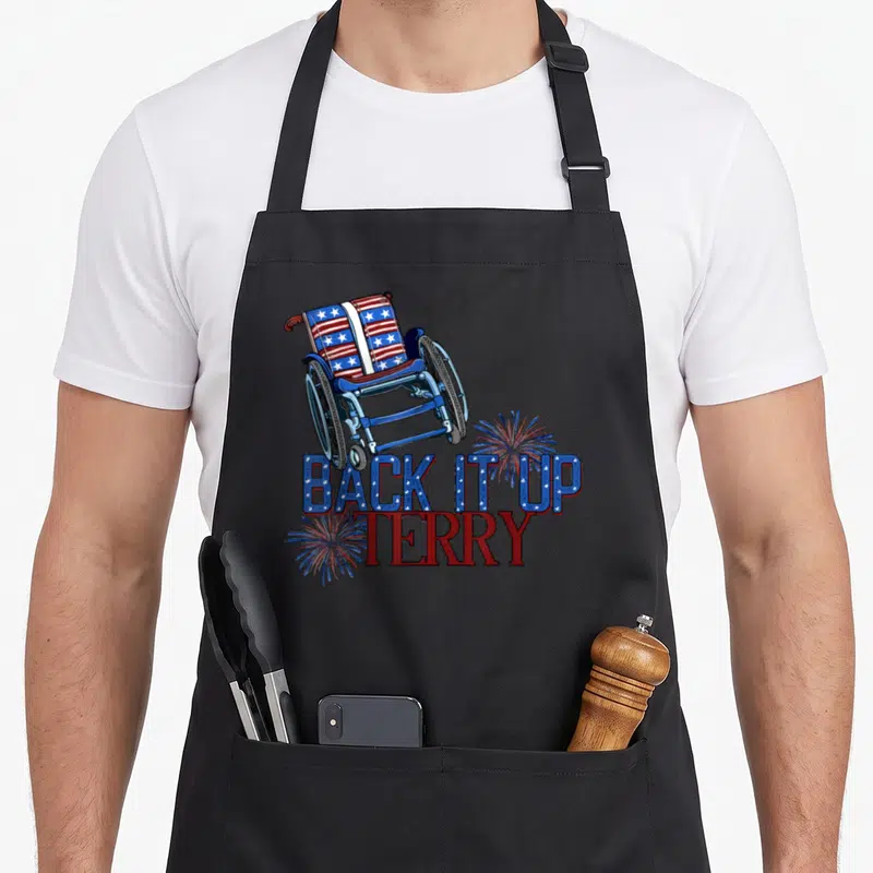 Terry reverse firework 4th of july apron - Wheelchair with flag pattern and BACK IT UP TERRY text surrounded by red — shown on a black cotton canvas apron against a white background