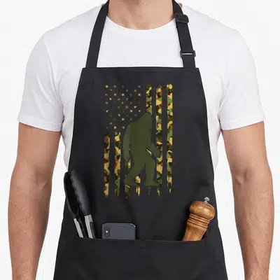 Foot camo aprons - Bigfoot silhouette against olive tan brown camouflage flag pattern with distressed texture — shown on a black cotton canvas apron against a white background