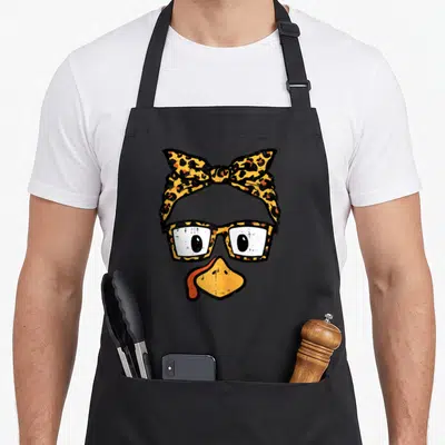 Face funny turkey apron - Cartoon duck face with leopard print glasses and bow headband in orange and black — shown on a black cotton canvas apron against a white background