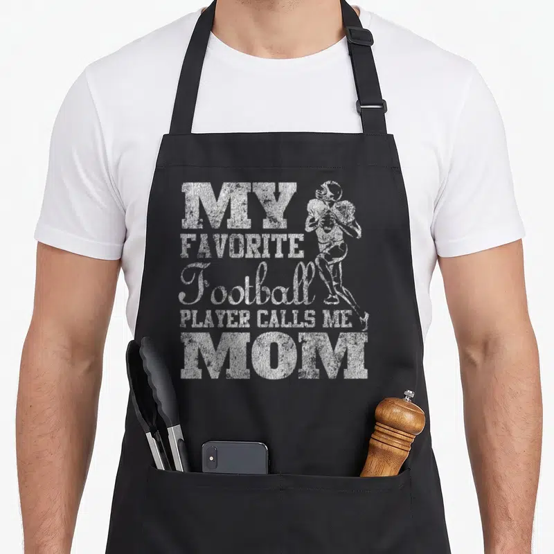 Player call football apron - Distressed white text reading MY FAVORITE Football PLAYER CALLS ME MOM next to a running — shown on a black cotton canvas apron against a white background