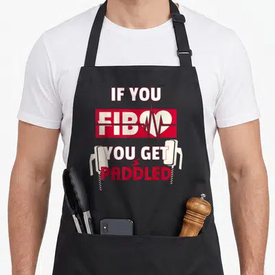Paramedic nurse apron - IF YOU FIB YOU GET PADDLED text with heart EKG line and two defibrillator paddles — shown on a black cotton canvas apron against a white background