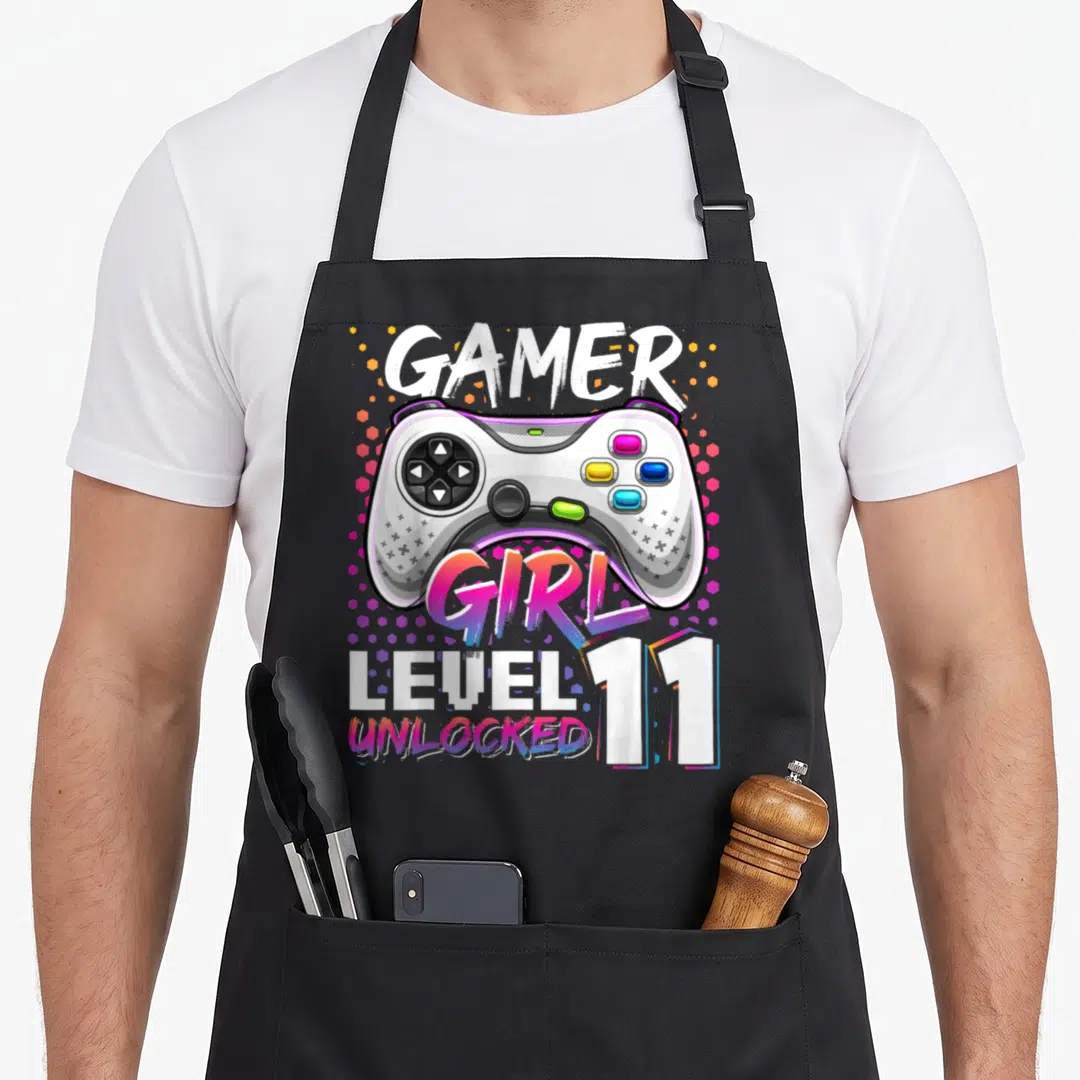 Gamer level unlocked video game apron - Cartoon controller with GAMER GIRL LEVEL 11 UNLOCKED text and colorful dots — shown on a black cotton canvas apron against a white background