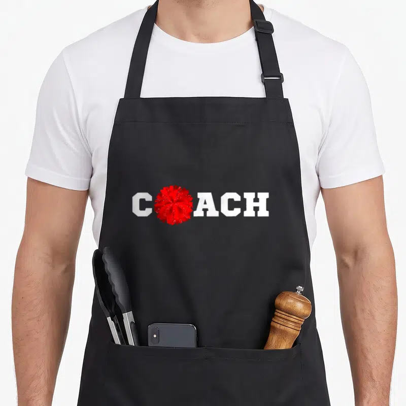 Cheer coach squad apron - White COACH text with red glitter pom-pom replacing the O letter — shown on a black cotton canvas apron against a white background