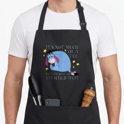 Much eeyore apron - Blue cartoon donkey holding tail with scattered yellow stars and split text quote — shown on a black cotton canvas apron against a white background