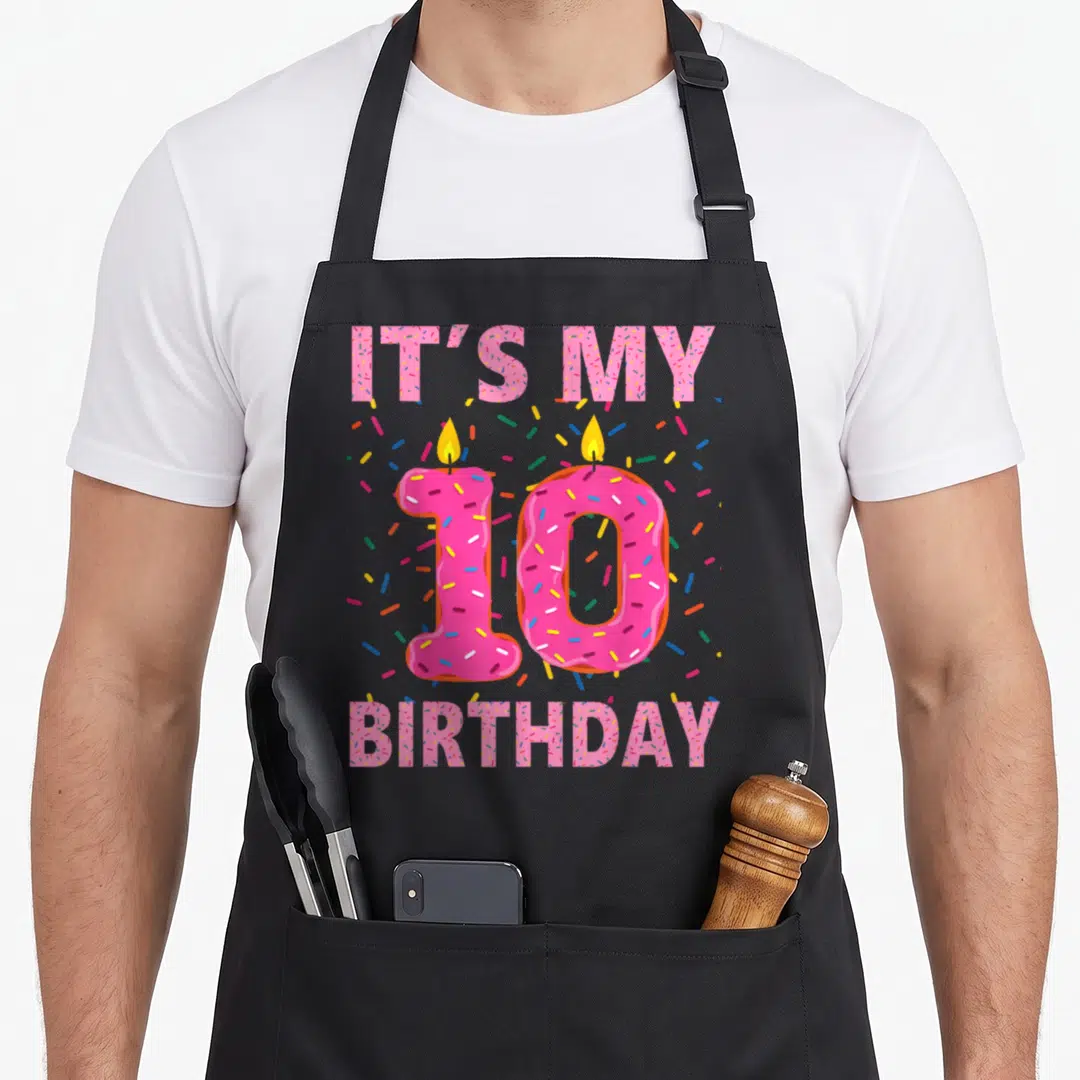 10th donut apron - Pink donut number 10 with candles and IT'S MY BIRTHDAY text surrounded by sprinkles — shown on a black cotton canvas apron against a white background