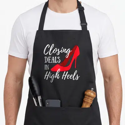 Closing real estate agent apron - Red high heel shoe next to white stacked text Closing Deals In High Heels — shown on a black cotton canvas apron against a white background