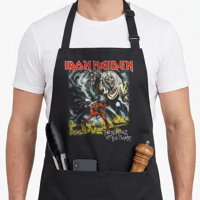 Eddie boy number apron - Iron Maiden text above skeletal monster and small red demon in grey and yellow — shown on a black cotton canvas apron against a white background