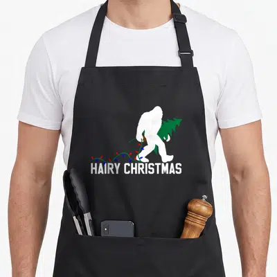 Hairy bigfoot apron - White Bigfoot silhouette carrying green tree and colorful lights above HAIRY CHRISTMAS text — shown on a black cotton canvas apron against a white background