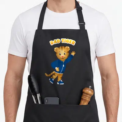 Daniel tiger dad apron - Arched DAD TIGER text above a waving cartoon tiger in a blue jacket — shown on a black cotton canvas apron against a white background