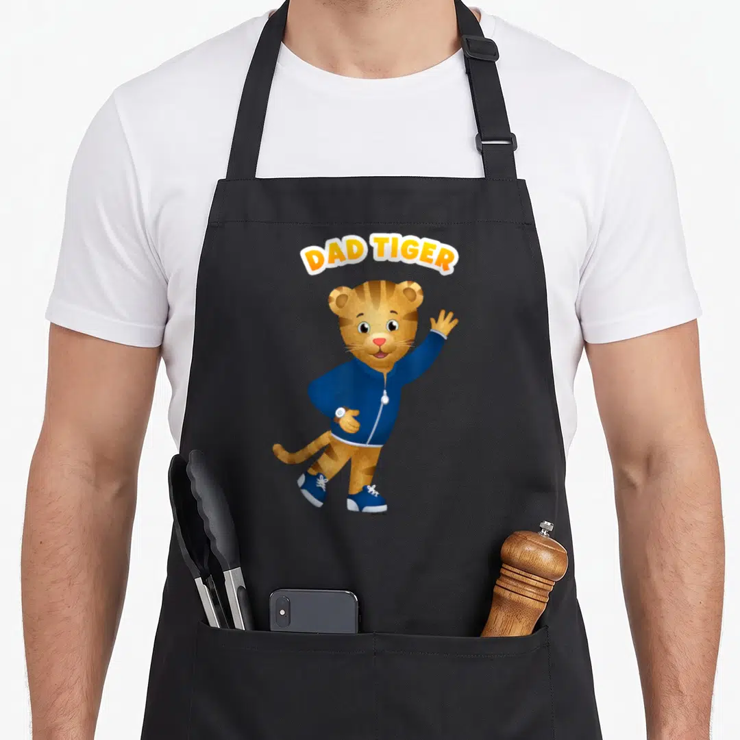 Daniel tiger dad apron - Arched DAD TIGER text above a waving cartoon tiger in a blue jacket — shown on a black cotton canvas apron against a white background