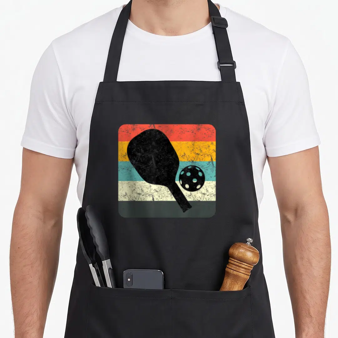 Silhouette pickleball apron - Black pickleball paddle and perforated ball over horizontal red, orange, teal, cream — shown on a black cotton canvas apron against a white background