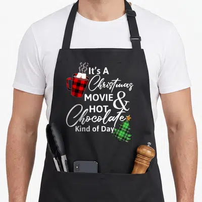 Hot christmas apron - Plaid hot chocolate mug and plaid tree with white text It's A Christmas MOVIE & Hot Chocolate — shown on a black cotton canvas apron against a white background