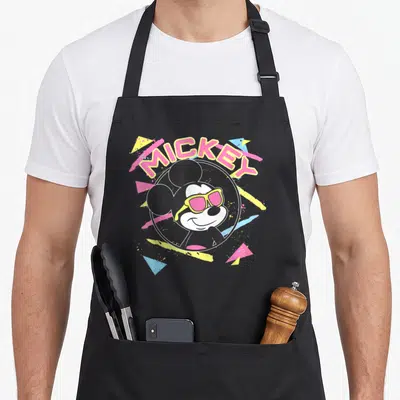 90s best friend aprons - Retro Mickey Mouse with pink sunglasses inside a circle with MICKEY text and geometric — shown on a black cotton canvas apron against a white background