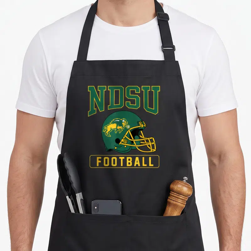 North football apron - Green football helmet with yellow bison and NDSU FOOTBALL text in cartoon style — shown on a black cotton canvas apron against a white background
