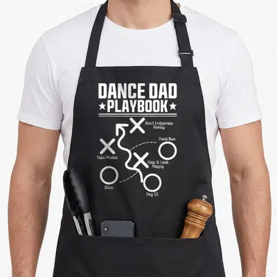 Comp daddy apron - White playbook diagram with Xs Os arrows and text labels for dance dad tasks — shown on a black cotton canvas apron against a white background