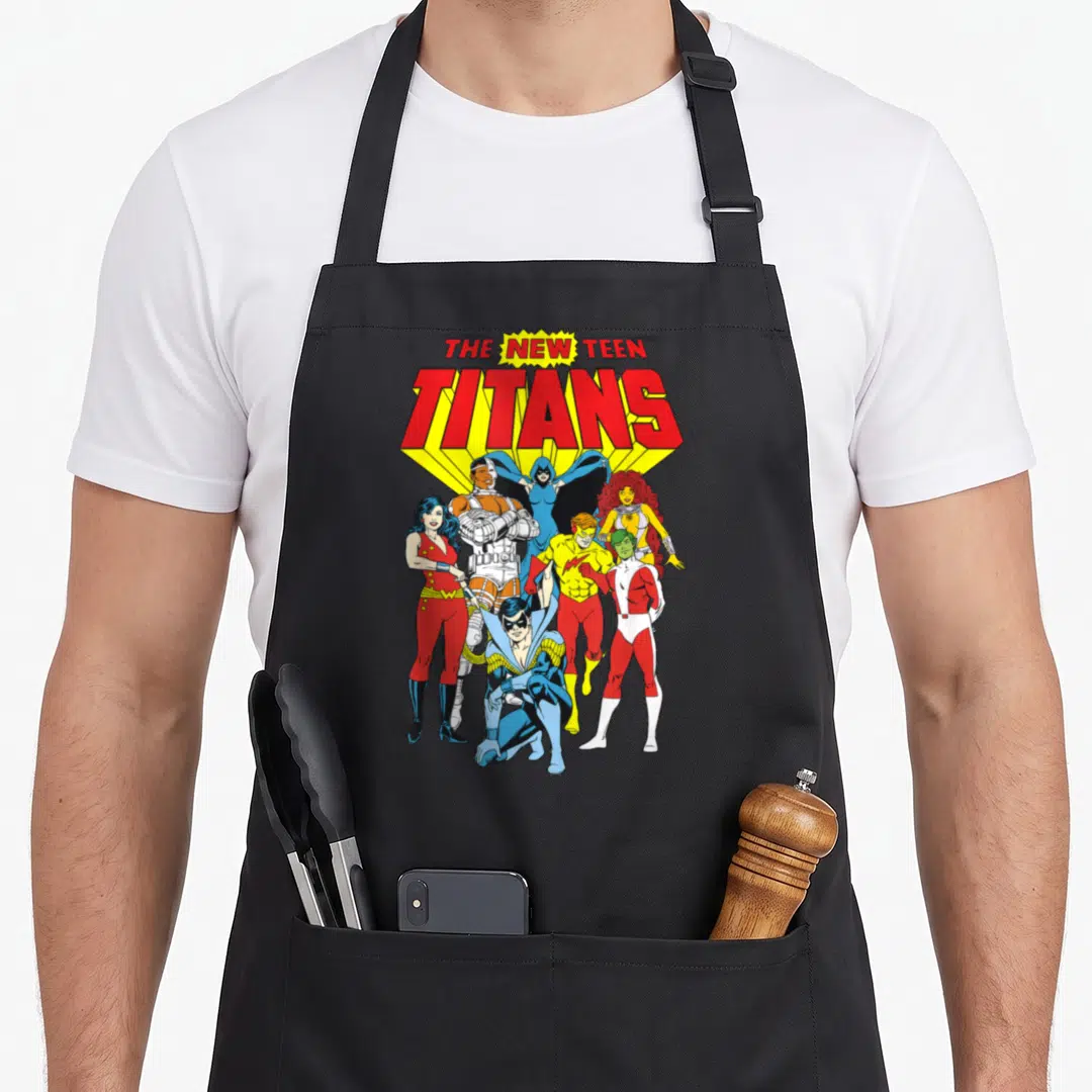 Justice novelty apron - Stacked THE NEW TEEN TITANS text above seven colorful superhero characters in cartoon poses — shown on a black cotton canvas apron against a white background
