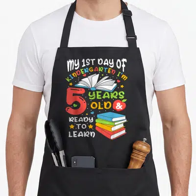 School kindergarten back apron - Cartoon text My 1st Day of Kindergarten with red 5, open book, apple, and stacked books — shown on a black cotton canvas apron against a white background