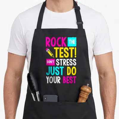 Stress test kitchen apron - Stacked pink cyan yellow white text reading ROCK THE TEST with a pencil graphic — shown on a black cotton canvas apron against a white background