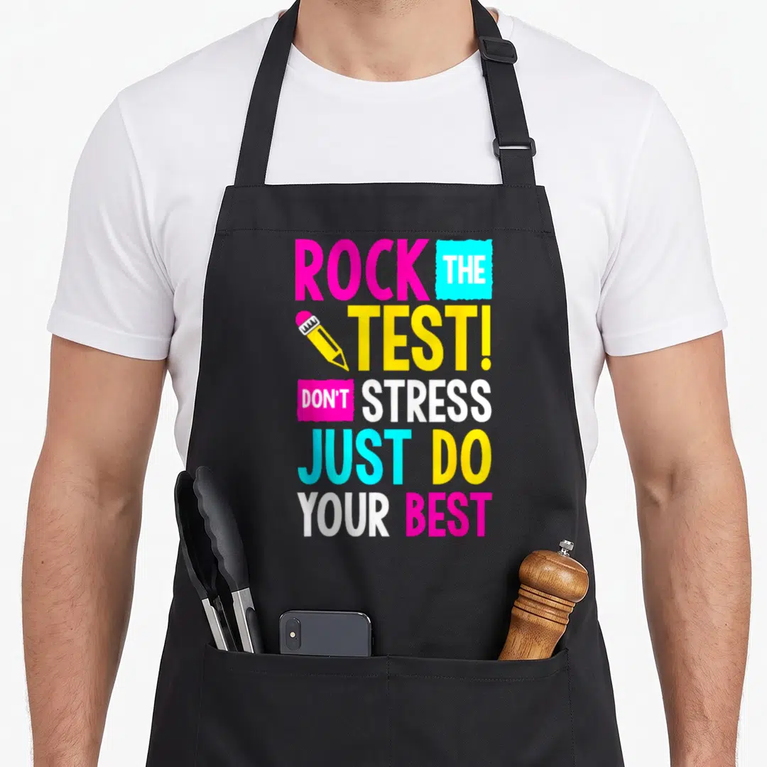 Stress test kitchen apron - Stacked pink cyan yellow white text reading ROCK THE TEST with a pencil graphic — shown on a black cotton canvas apron against a white background