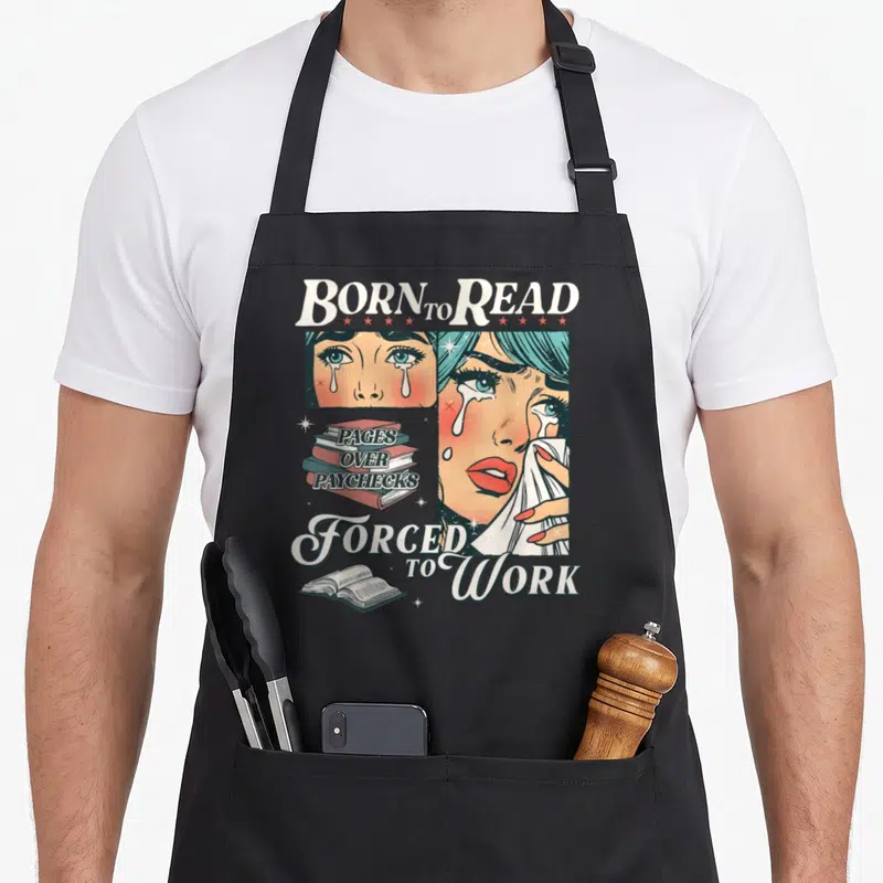 Read book lovers apron - BORN TO READ text above crying cartoon woman and stack of books reading PAGES OVER PAYCHECKS — shown on a black cotton canvas apron against a white background