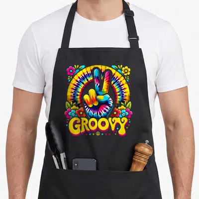 Sign tie-dye apron - Tie-dye hand making a peace sign with groovy text and flowers in yellow pink blue and green — shown on a black cotton canvas apron against a white background