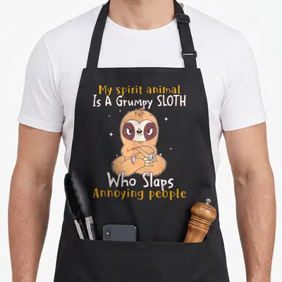 Slap animal apron - Grumpy cartoon sloth with crossed arms and text about slapping annoying people — shown on a black cotton canvas apron against a white background