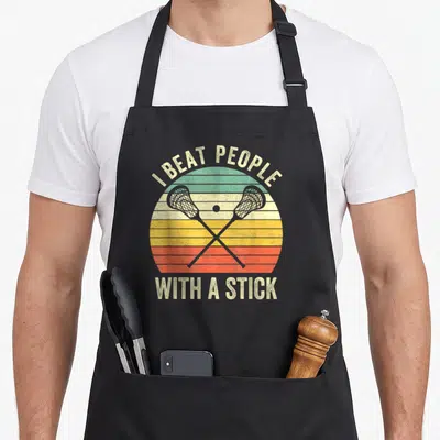 Beat lacrosse apron - Crossed lacrosse sticks over a striped sunset with the text I BEAT PEOPLE WITH A STICK — shown on a black cotton canvas apron against a white background