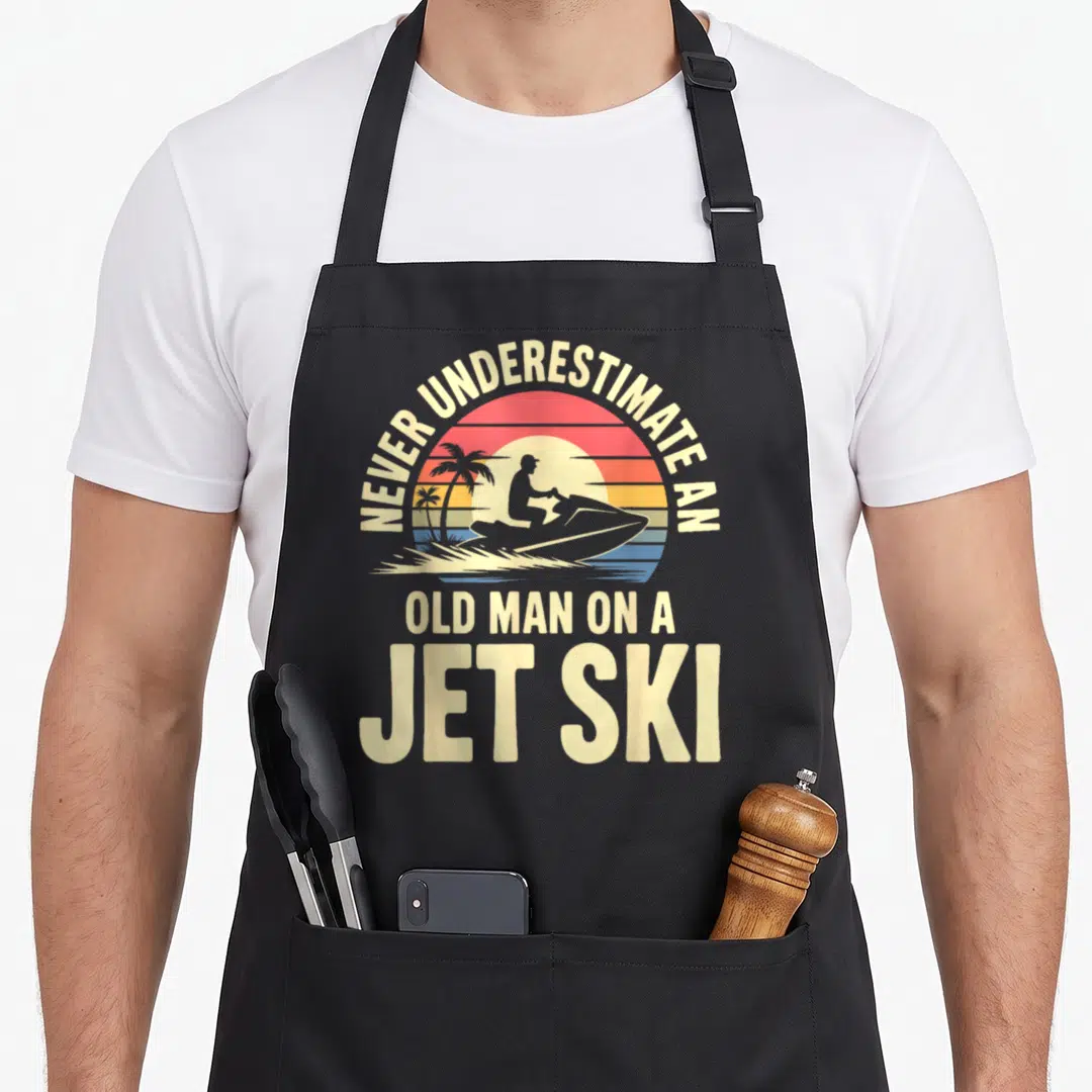 Jet grandpa apron - Retro sun graphic with jet ski silhouette and cream text reading Never Underestimate An Old Man — shown on a black cotton canvas apron against a white background