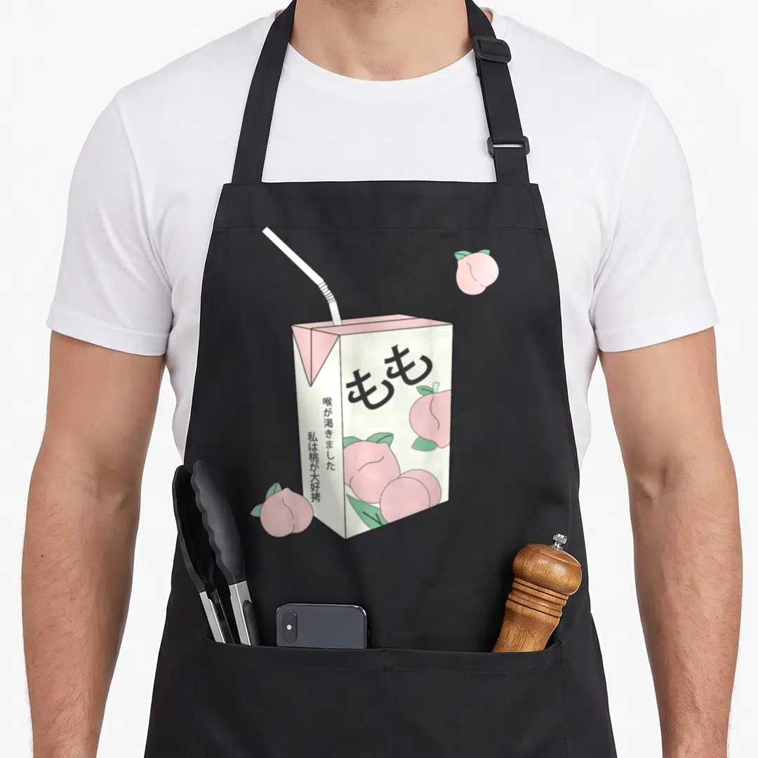 Japanese indie apron - Pink peach juice box with straw, Japanese text, and scattered peaches in cartoon style — shown on a black cotton canvas apron against a white background