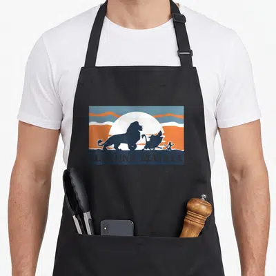 Matata lion apron - Retro lion warthog meerkat silhouettes under white sun with Hakuna Matata text — shown on a black cotton canvas apron against a white background