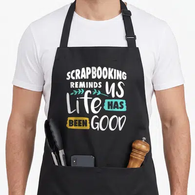 Scrapbooking us aprons - White and colored text reading Life Has Been Good with teal leaves on black fabric — shown on a black cotton canvas apron against a white background