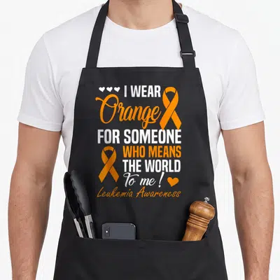 Someone cancer awareness apron - I Wear Orange stacked text with two orange ribbons and white and orange hearts — shown on a black cotton canvas apron against a white background
