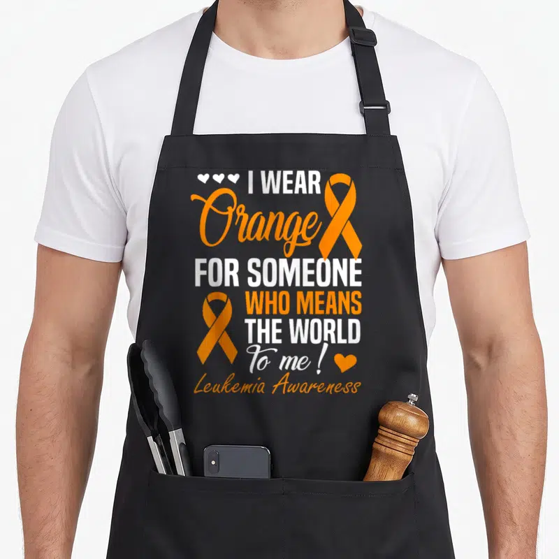 Someone cancer awareness apron - I Wear Orange stacked text with two orange ribbons and white and orange hearts — shown on a black cotton canvas apron against a white background