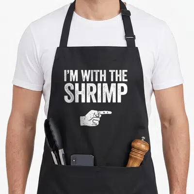 Costume shrimp apron - Distressed white text I'M WITH THE SHRIMP above a pointing hand on black apron — shown on a black cotton canvas apron against a white background
