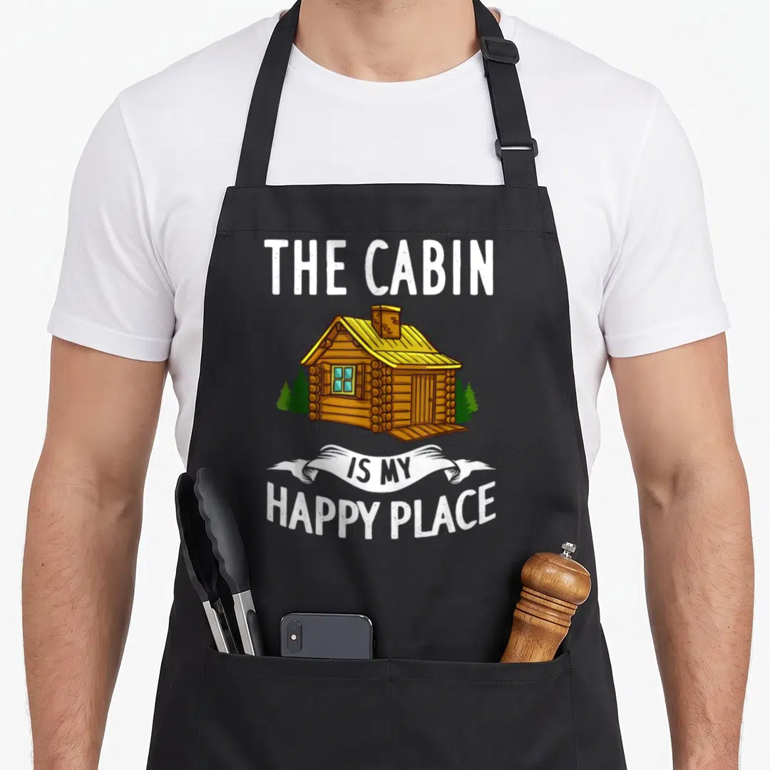 Berghaus cabin apron - Cartoon log cabin with yellow roof and green trees under text reading The Cabin Is My Happy — shown on a black cotton canvas apron against a white background