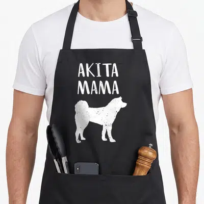 Pet inu dog apron - White distressed text reading AKITA MAMA above a standing dog silhouette — shown on a black cotton canvas apron against a white background