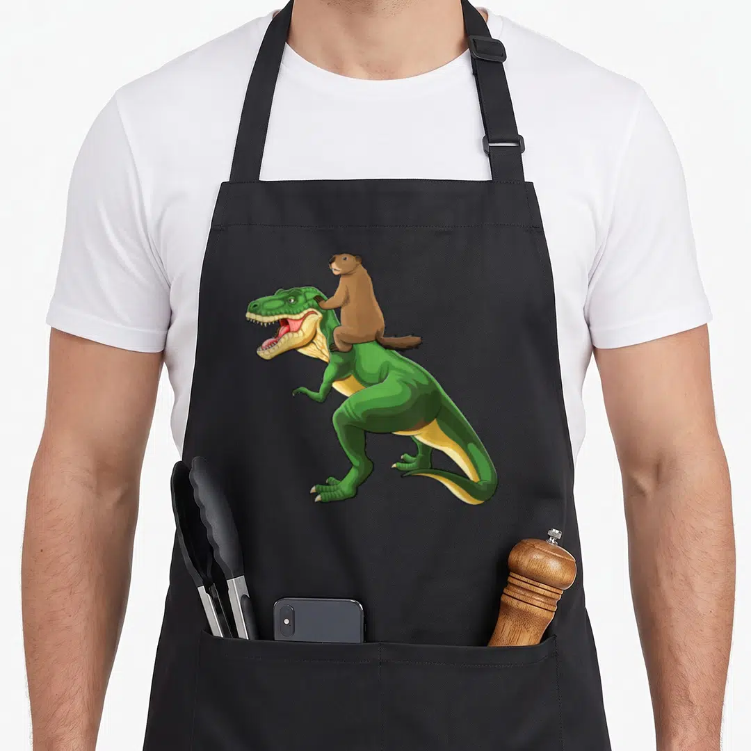 Groundhog dinosaur apron - Green T-Rex with brown beaver riding on back in cartoon style — shown on a black cotton canvas apron against a white background