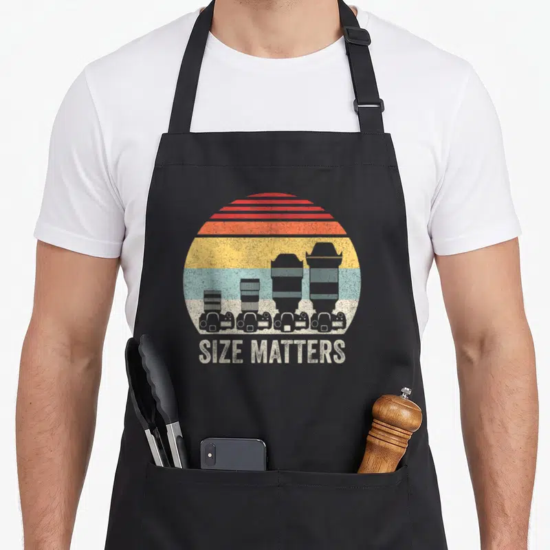 Catch retro camera apron - Four cameras with growing lenses under a striped sun with SIZE MATTERS text — shown on a black cotton canvas apron against a white background