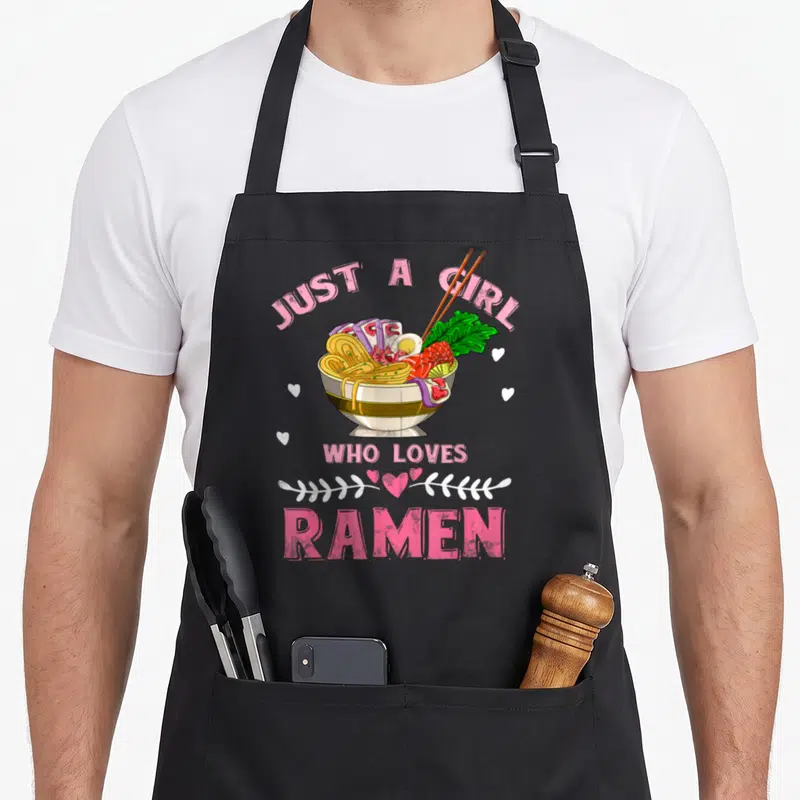 Japanese food anime apron - Cartoon ramen bowl with chopsticks and pink text reading JUST A GIRL WHO LOVES RAMEN — shown on a black cotton canvas apron against a white background