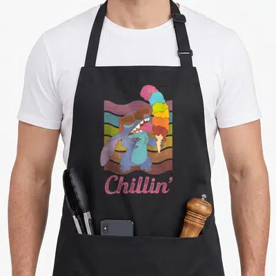 Rainbow ice cream apron - Blue cartoon character holding multi-colored ice cream cone against wavy rainbow stripes — shown on a black cotton canvas apron against a white background