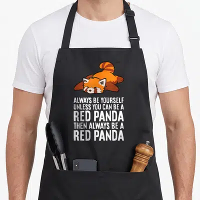 Panda four raccoon apron - Lying cartoon red panda above five lines of white text reading Always Be Yourself Unless You — shown on a black cotton canvas apron against a white background