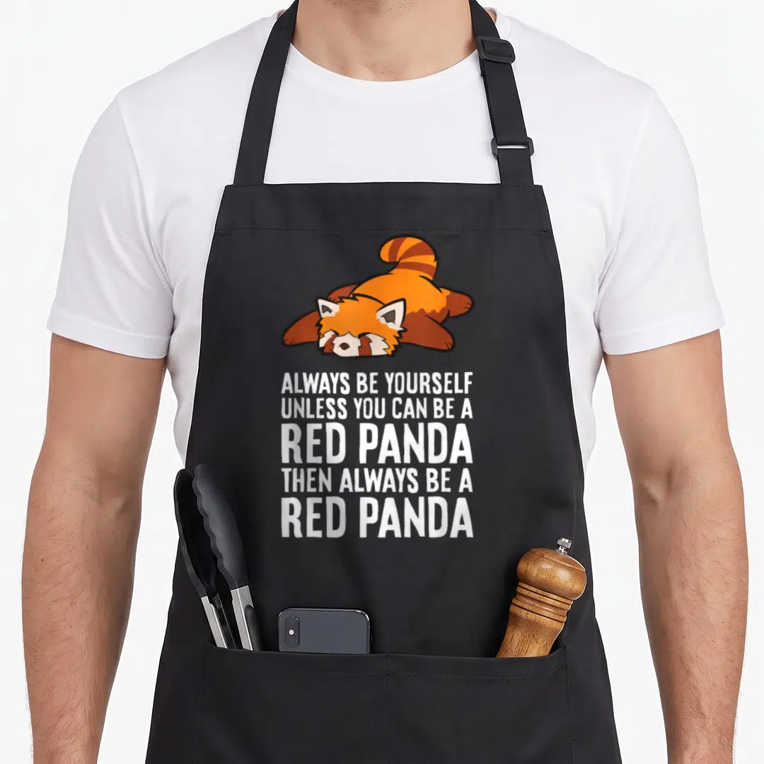 Panda four raccoon apron - Lying cartoon red panda above five lines of white text reading Always Be Yourself Unless You — shown on a black cotton canvas apron against a white background