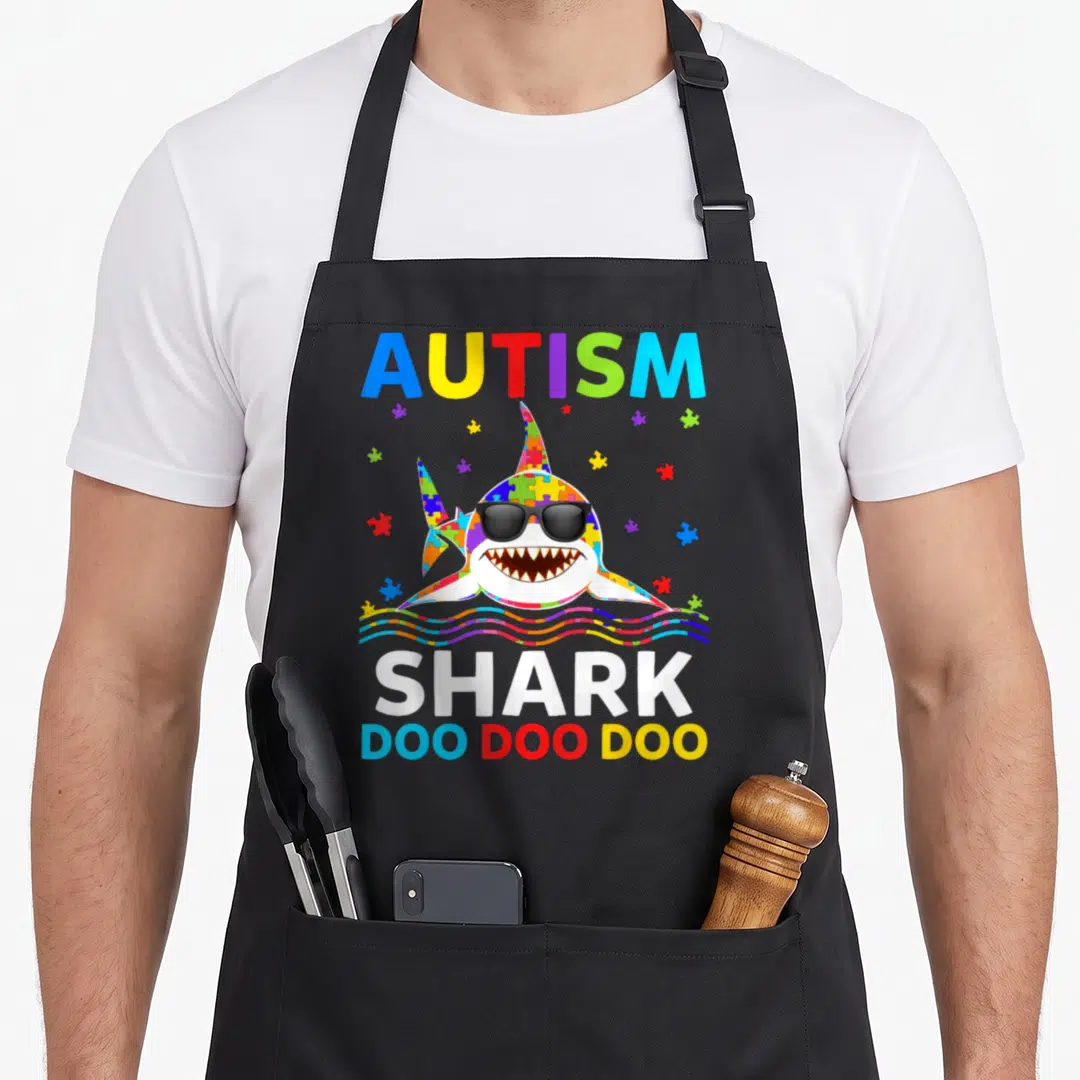Autism shark dad apron - Cartoon puzzle-piece shark with sunglasses and rainbow waves under AUTISM text — shown on a black cotton canvas apron against a white background
