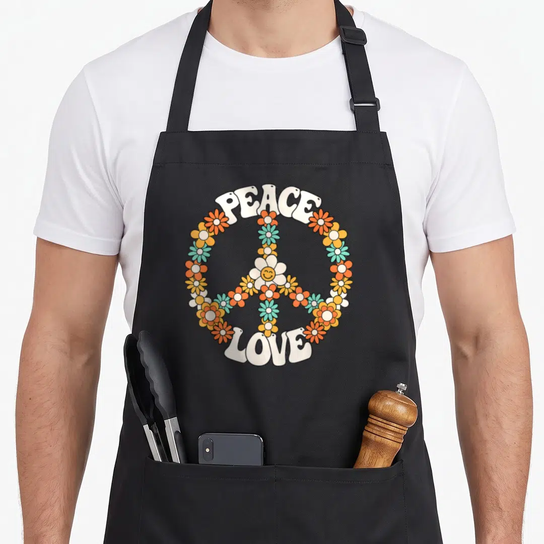 Outfit 60s apron - Floral peace sign with arched PEACE and LOVE text in cartoon style — shown on a black cotton canvas apron against a white background