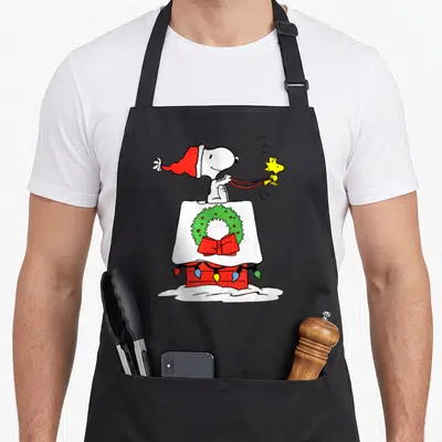 Doghouse holiday apron - Snoopy in red hat behind snow-covered doghouse with green wreath and yellow lights holding — shown on a black cotton canvas apron against a white background
