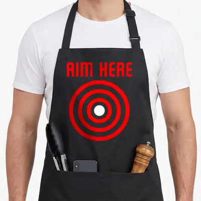 Bullseye target apron - Red concentric circles with white center and red AIM HERE text above — shown on a black cotton canvas apron against a white background