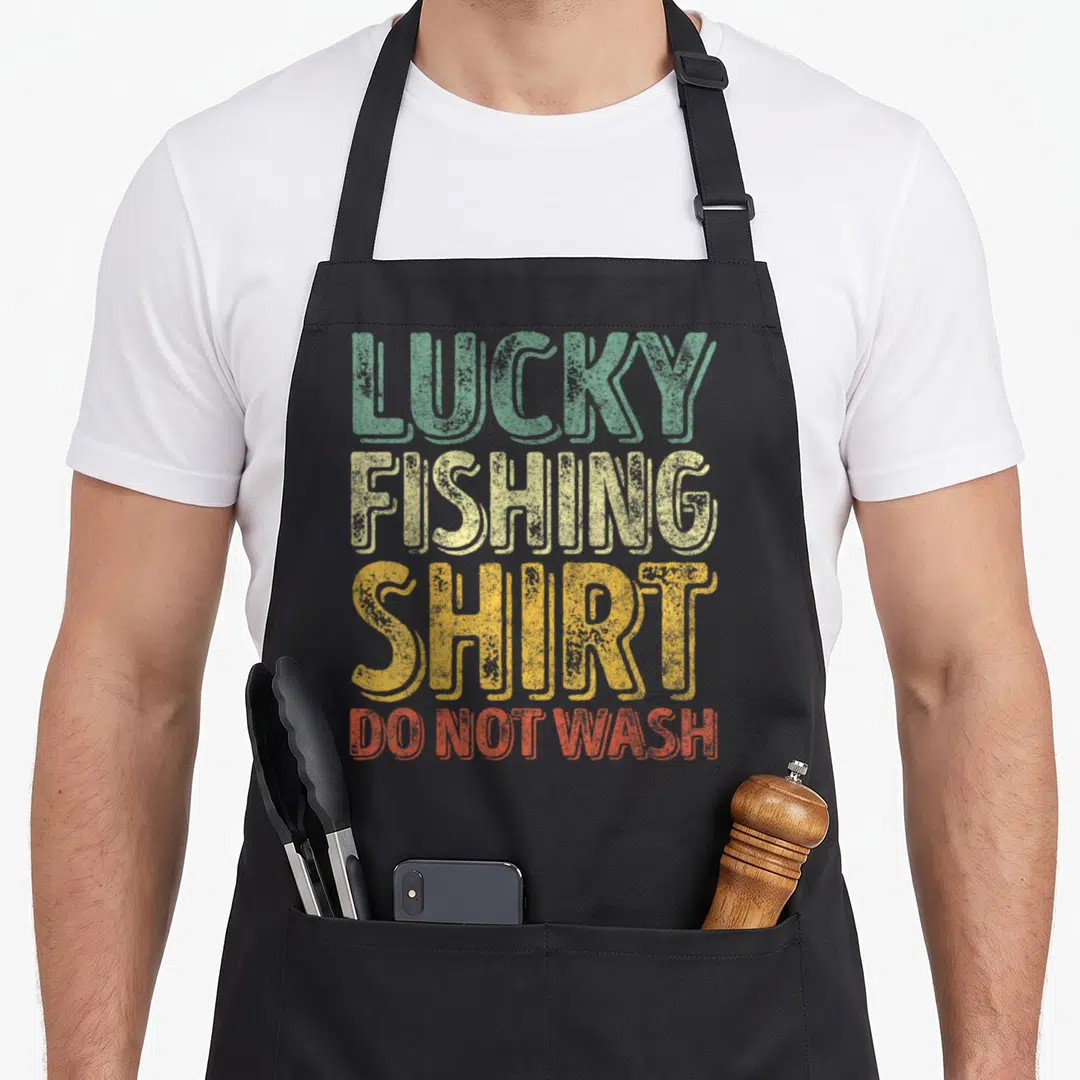 Lucky fisherman apron - Stacked distressed text reading LUCKY FISHING SHIRT DO NOT WASH in green yellow and red — shown on a black cotton canvas apron against a white background