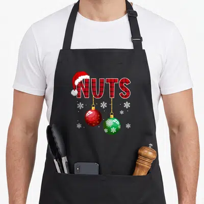Funny chest couples christmas aprons - NUTS text in red plaid with Santa hat, hanging red and green baubles, and white — shown on a black cotton canvas apron against a white background