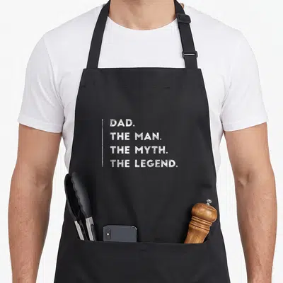 Daughter birthday apron - White stacked text DAD THE MAN THE MYTH THE LEGEND with vertical line — shown on a black cotton canvas apron against a white background