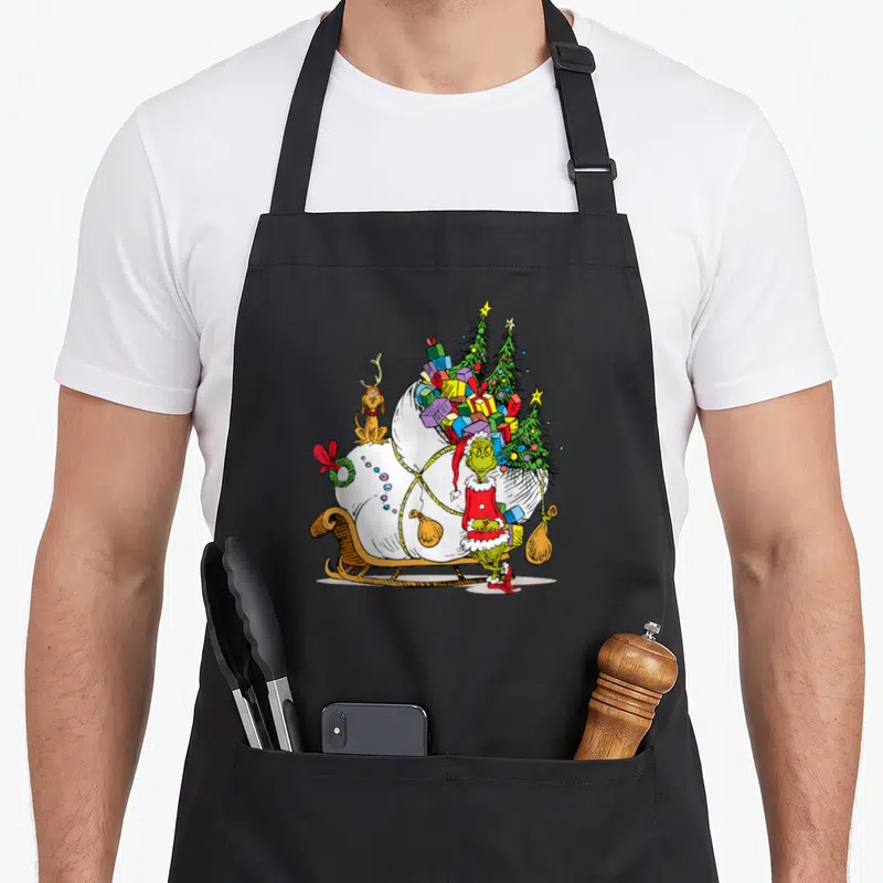 Max holiday apron - Grinch in red suit standing next to Max on a sleigh loaded with a white sack of gifts — shown on a black cotton canvas apron against a white background